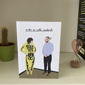 Puede incluir: Una tarjeta de felicitaci&oacute;n con una ilustraci&oacute;n de dibujos animados de dos personas. Una persona lleva un disfraz de esqueleto amarillo y la otra lleva una camisa azul y pantalones grises. El texto de la tarjeta dice "make an exotic sandwich".