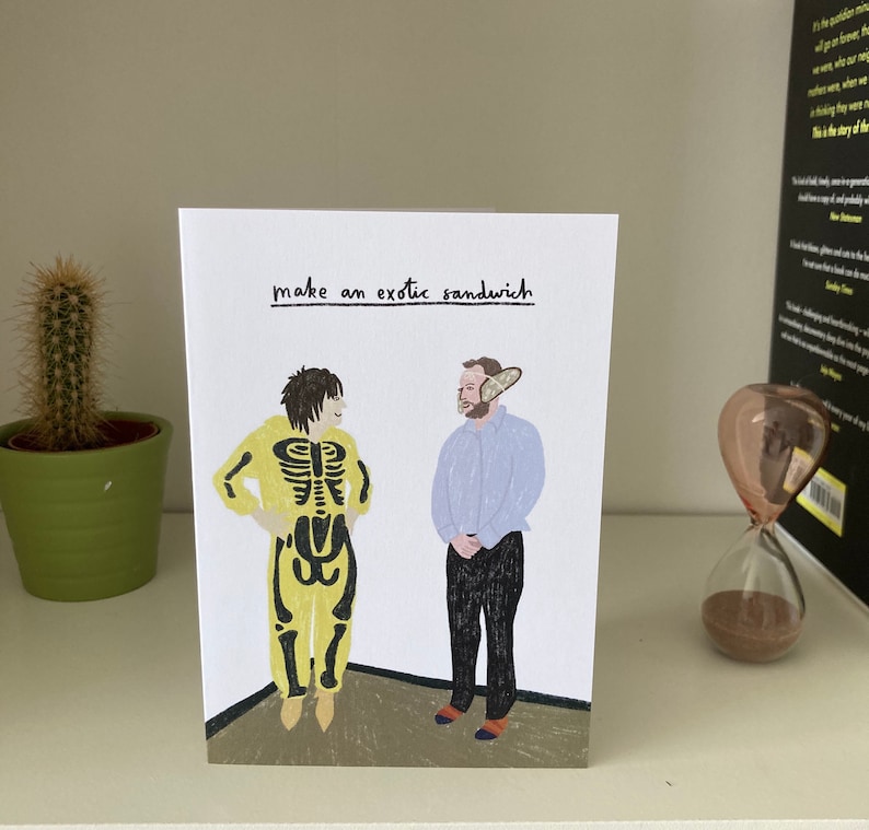 Puede incluir: Una tarjeta de felicitaci&oacute;n con una ilustraci&oacute;n de dibujos animados de dos personas. Una persona lleva un disfraz de esqueleto amarillo y la otra lleva una camisa azul y pantalones grises. El texto de la tarjeta dice "make an exotic sandwich".