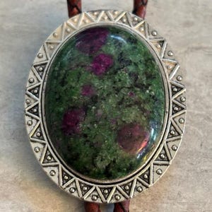 May include: A silver bolo tie with a green and pink oval stone set in a decorative silver frame. The bolo tie has brown leather cords.