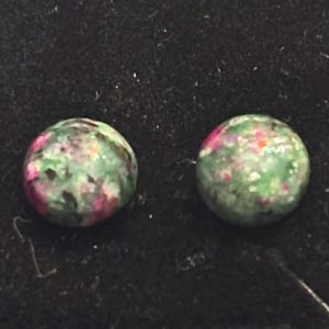 May include: A pair of green and pink gemstone earrings. The earrings are round and have a speckled pattern.