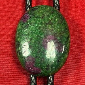 May include: A green and purple gemstone bolo tie with a black braided cord.