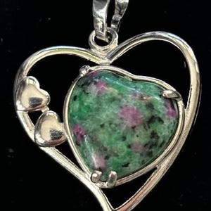 May include: A silver heart-shaped pendant with a green and pink gemstone in the center. The pendant has three smaller hearts on the side.