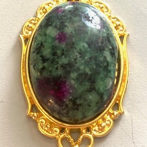 May include: A gold-colored pendant with an oval-shaped, green and purple stone. The stone is set in a decorative gold frame with ornate detailing. The pendant has a loop at the top for a chain.