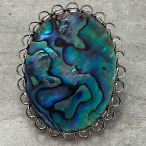 May include: A silver oval-shaped brooch with a large iridescent abalone shell inlay. The shell has a swirling pattern of blue, green, and black.