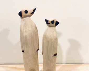 May include: Two standing meerkat figurines carved from light-colored wood. Each figure features dark, burnt-effect detailing on the ears, eyes, nose, and feet, with a smooth, natural wood grain finish visible across the elongated, upright bodies.