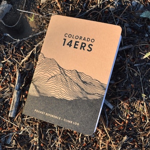 Może przedstawiać: Brązowo-czarny notatnik kieszonkowy z napisem "COLORADO 14ERS" i grafiką gór. Notatnik leży na posłaniu z igieł sosnowych i gałązek. Na dole notatnika widnieje napis "POCKET REFERENCE - CLIMB LOG".
