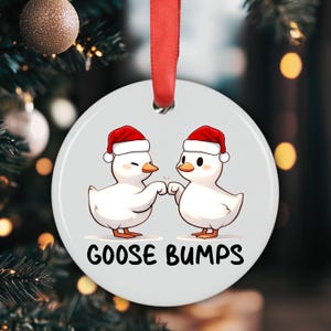 May include: A white ceramic Christmas ornament with a red ribbon for hanging. The ornament features two cartoon ducks wearing Santa hats, bumping beaks, and the text "GOOSE BUMPS" in black.
