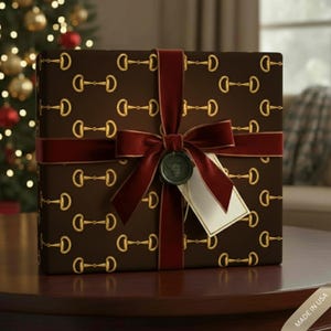 May include: A wrapped gift box with a brown background and gold horse bit pattern. It is tied with a burgundy velvet ribbon and bow, and a small tag hangs from the bow. The gift is on a wooden table, with a Christmas tree in the background.