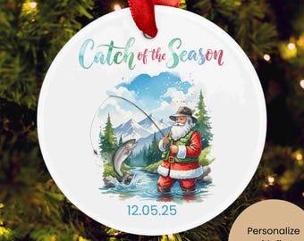 Puede incluir: Adorno navideño redondo y blanco con una cinta roja. El adorno presenta a Papá Noel pescando en un río, con un fondo de montaña. El texto dice "Catch of the Season" y "12.05.25".