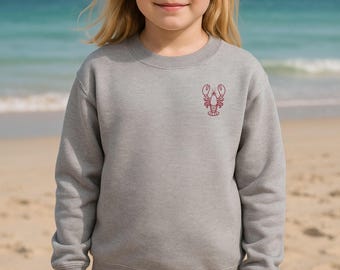 Könnte beinhalten: Graues Sweatshirt mit roter Hummer-Grafik auf der Brust. Das Sweatshirt besteht aus einem weichen Material und hat einen Rundhalsausschnitt. Der Hintergrund zeigt einen Strand mit Sand und Meer.