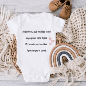 May include: White baby onesie with a sweet message in Spanish: "Mi pequeña, ¡qué orgulloso estoy! Mi pequeña, no te dejaré. Mi pequeña, yo te cuidaré. Y por siempre te amaré." The onesie is surrounded by a woven basket, a crocheted blanket, and a rainbow-colored macrame.