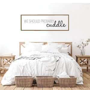 May include: A white bedroom with a wooden bed frame and white bedding. The bed has a sign above it that reads "We should probably cuddle".