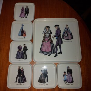 May include: Set of 8 white ceramic coasters with colorful illustrations of people in traditional clothing from different regions of Germany. Each coaster features a different region and year, including:  "Trachtenvogesen 1911", "Schwäbisch Alb 1911", "Leutkirch 1900", "Allgäu 1907", "Grünberg 1907", "Wildberg 1939", "Hechingen 1920".