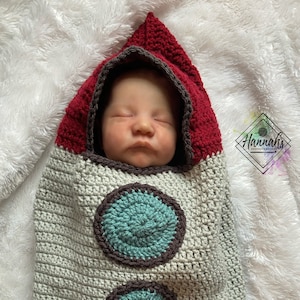 May include: A baby swaddled in a crocheted rocket-shaped sleeping sack. The sack is gray with brown and red accents and features two teal circles. The baby is wearing a red crocheted hat.
