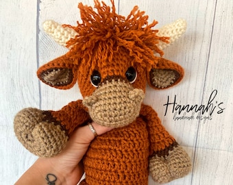 May include: A crocheted stuffed toy Highland cow with brown and tan fur, large brown eyes, and a white nose. The toy is being held by a hand with a tattoo.