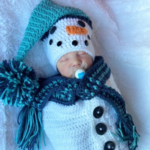 Op de afbeelding: Een wit gebreid sneeuwmanpak met een blauwe en groene muts en sjaal. Het pak heeft drie zwarte knopen en een pompon op de muts. Het pak ligt op een wit pluche oppervlak. Hannah's Handmade Designs.