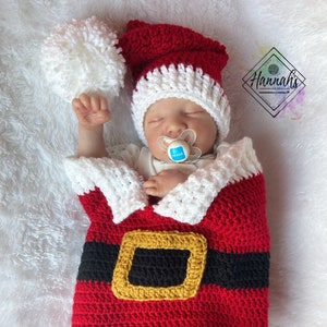 Op de afbeelding: Een rood en wit gebreid kerstmanpakje voor een baby, met een bijpassende muts en een witte pompon. Het pakje heeft een zwarte riem met een gouden gesp.