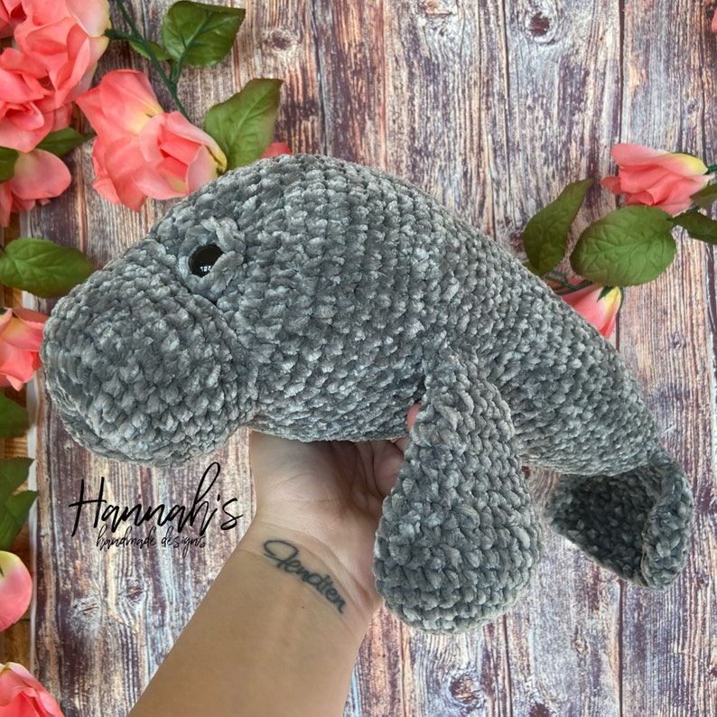 Puede incluir: Un juguete de peluche de manat&iacute; de crochet gris. El juguete est&aacute; siendo sostenido por una mano con un tatuaje que dice "Jenction".