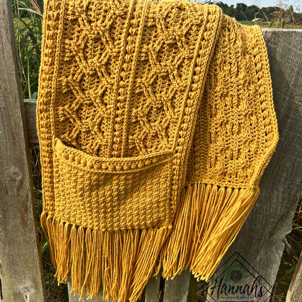 May include: A mustard yellow crocheted scarf with a pocket and fringe. The scarf is hanging on a wooden fence.