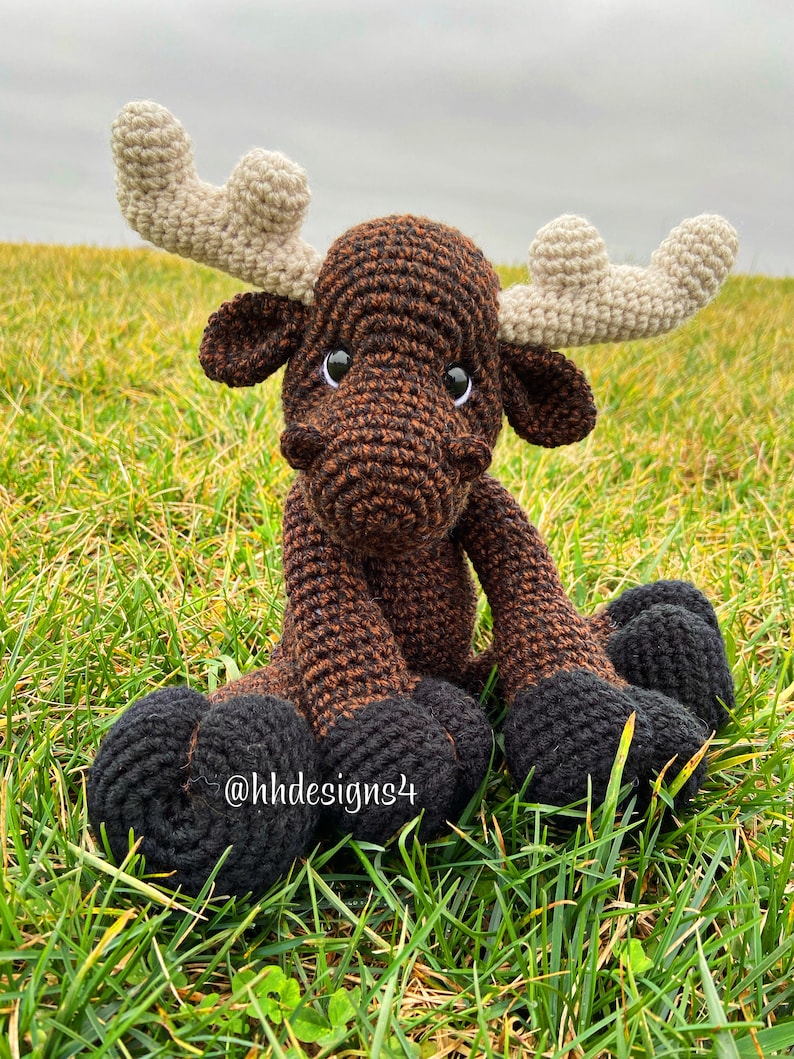 Puede incluir: Un alce de crochet marr&oacute;n y negro con grandes cuernos. El alce est&aacute; sentado en un lecho de hierba verde. El alce tiene ojos negros y una nariz marr&oacute;n. El texto '@hhdesigns4' es visible en la pierna del alce.