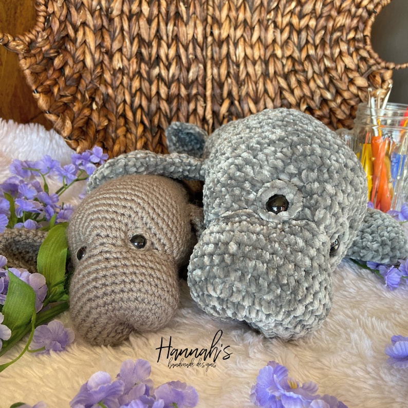 Puede incluir: Dos peluches de manat&iacute; de crochet, uno gris y otro marr&oacute;n, est&aacute;n tumbados sobre una superficie blanca y esponjosa con flores moradas. El manat&iacute; gris tiene una superficie texturizada y peluda. El manat&iacute; marr&oacute;n tiene una superficie lisa. Los peluches est&aacute;n hechos a mano y tienen ojos negros.