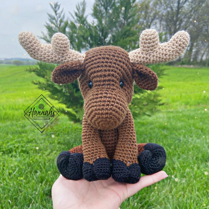 Puede incluir: Un alce de crochet marr&oacute;n con pezu&ntilde;as negras y grandes cuernos beige. El alce est&aacute; sentado en una mano.