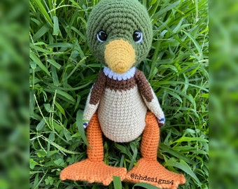 May include: A crocheted green duck with orange feet and a white beak. The duck is sitting in a bed of green grass.