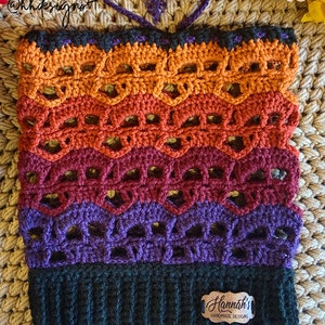 Puede incluir: Un gorro de crochet con un degradado de color negro, morado, rojo, naranja y amarillo. El gorro tiene un patrón de encaje y un borde negro. El gorro tiene una etiqueta que dice "Hannah's Handmade Designs".