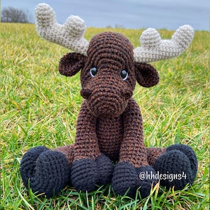 Puede incluir: Un alce de crochet marr&oacute;n y negro con grandes cuernos blancos. El alce est&aacute; sentado en un lecho de hierba verde. El alce tiene ojos negros y una nariz marr&oacute;n. El alce es un juguete hecho a mano.