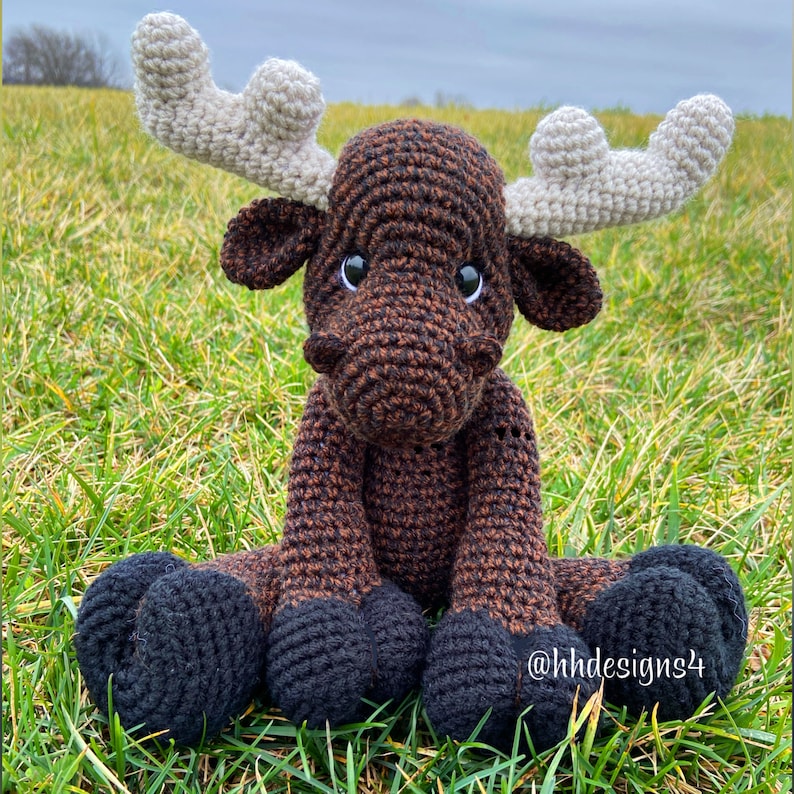 Puede incluir: Un alce de crochet marr&oacute;n y negro con grandes cuernos blancos. El alce est&aacute; sentado en un lecho de hierba verde. El alce tiene ojos negros y una nariz marr&oacute;n. El alce es un juguete hecho a mano.