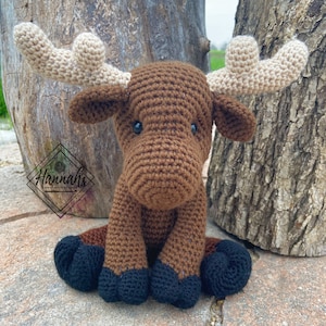 Puede incluir: Un alce de crochet marr&oacute;n con grandes cuernos beige. El alce est&aacute; sentado en una superficie marr&oacute;n con las patas metidas debajo del cuerpo. El alce tiene ojos negros y una nariz marr&oacute;n.