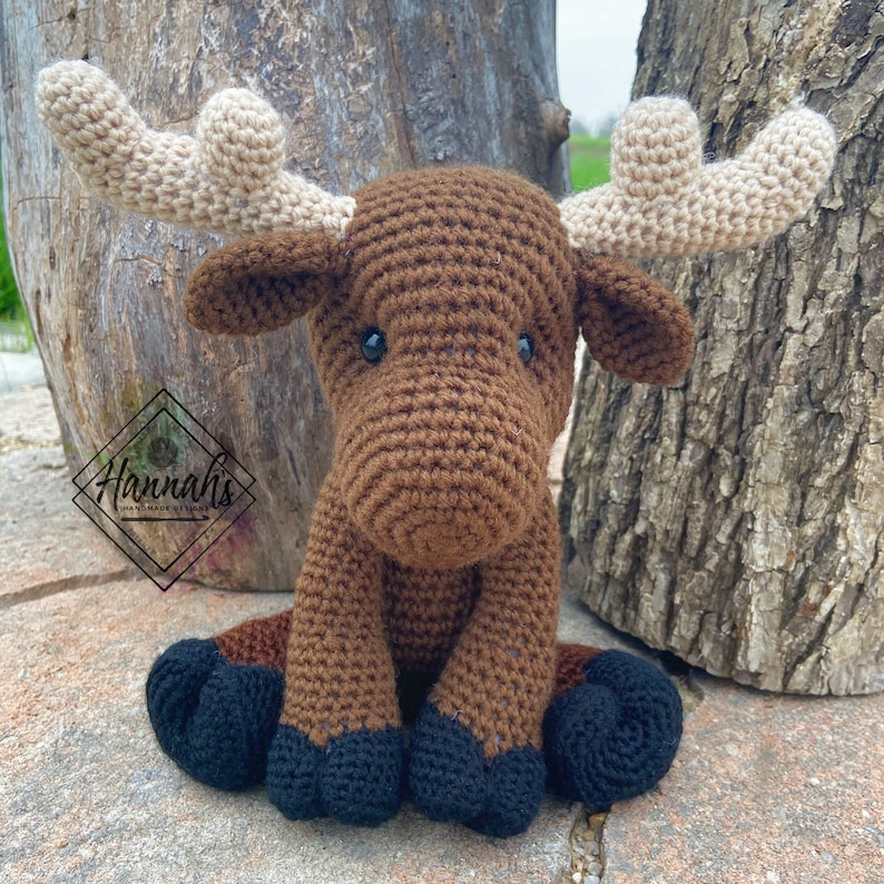 Puede incluir: Un alce de crochet marr&oacute;n con grandes cuernos beige. El alce est&aacute; sentado en una superficie marr&oacute;n con las patas metidas debajo del cuerpo. El alce tiene ojos negros y una nariz marr&oacute;n.