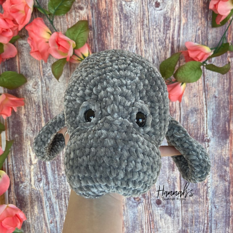 Puede incluir: Un juguete de peluche de manat&iacute; de crochet gris con ojos negros. El juguete se sostiene en una mano contra un fondo de madera con flores rosas.