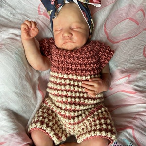 May include: A baby doll dressed in a handmade crochet outfit and a floral headband. The outfit is a mix of pink and cream. The headband is navy blue with a floral design. The doll rests on a pink and white patterned blanket.