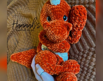 May include: A crocheted orange dinosaur toy with a white horn and white accents. The toy is sitting on a brown fuzzy surface.