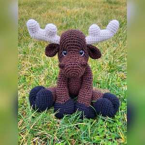 Puede incluir: Un juguete de alce de crochet marr&oacute;n y negro con cuernos blancos. El alce est&aacute; sentado en un lecho de hierba verde.