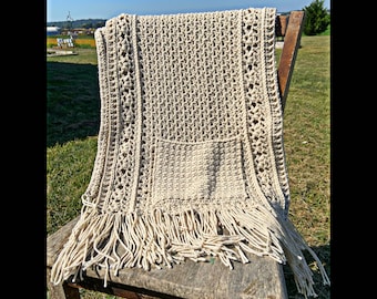 May include: A crocheted throw blanket with a fringe border in a natural beige colour. The blanket is draped over a wooden chair and has a pocket in the centre.