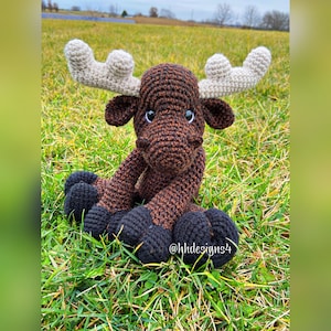 Puede incluir: Un alce de crochet marr&oacute;n y negro con cuernos blancos sentado en la hierba verde. El alce tiene ojos negros y una nariz marr&oacute;n. El texto "@hhdesigns4" es visible en la hierba.