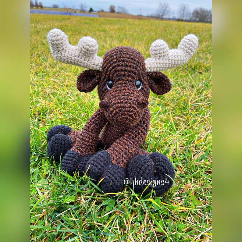 Puede incluir: Un alce de crochet marr&oacute;n y negro con cuernos blancos sentado en la hierba verde. El alce tiene ojos negros y una nariz marr&oacute;n. El texto "@hhdesigns4" es visible en la hierba.