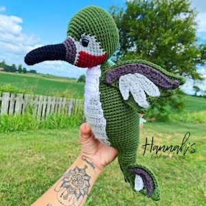 Pode incluir: Um brinquedo de pelúcia de beija-flor de crochê com detalhes em verde, cinza e branco. O brinquedo tem um bico longo e fino e uma garganta vermelha. O brinquedo está sendo segurado por uma mão com uma tatuagem.