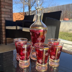 Puede incluir: Un juego de licor vintage que incluye una jarra con tapón y seis vasos de chupito a juego. El juego presenta un tono rojo rubí con adornos dorados y delicados grabados florales. La jarra y los vasos se exhiben sobre una superficie reflectante.