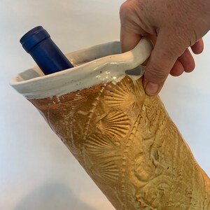 May include: A ceramic wine bottle cooler with a textured, embossed design. The cooler is a light brown color with a white rim and a handle. A blue wine bottle is partially inserted into the cooler.