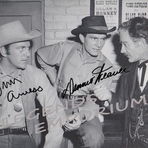 May include: Black and white photo of three men, two wearing cowboy hats. The men are standing in front of a brick wall with posters behind them. The posters have text that reads "$1,500 Reward William H. Bonney", "$2,000 Reward for Black Bart", and "$500 Reward Tommy B. Wanted for the Villa". The men are all smiling and looking at the camera. The photo is signed by two of the men, "Jim Amess" and "Dennis Weaver".