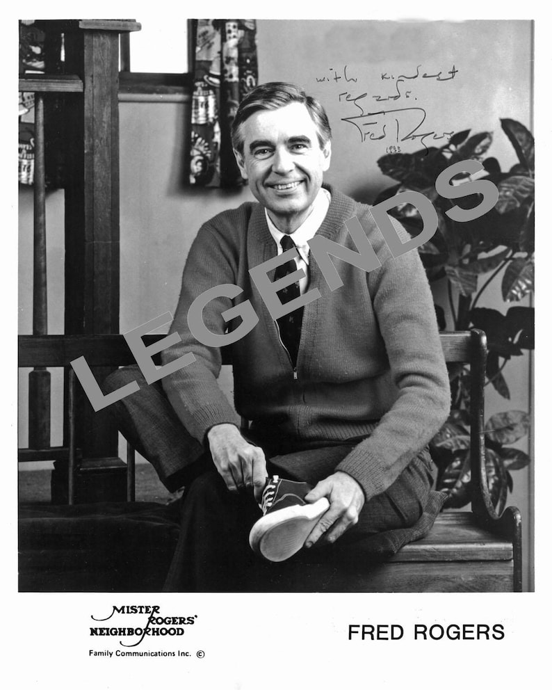 May include: Black and white photograph of Fred Rogers, seated and smiling, holding a small pair of sneakers. He wears a zip-up sweater, collared shirt, and tie. The image includes the text "Mister Rogers' Neighborhood" and "Fred Rogers".