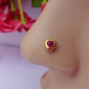 May include: A gold nose stud featuring a heart-shaped setting with a vibrant red gemstone at its center. The piece is designed with a smooth, polished metal frame that secures the faceted stone, creating a distinct heart silhouette on the skin.