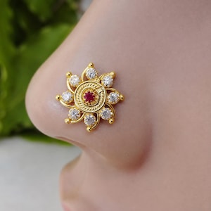 May include: A gold nose stud featuring a central circular red stone surrounded by a ring of eight clear white stones arranged in a floral pattern. The metal setting has a textured, rope-like border encircling the small, vibrant red center stone.