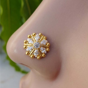 May include: A gold-colored nose stud featuring a floral design. The stud has a central, round, clear gemstone surrounded by smaller, teardrop-shaped clear gemstones. The design is accented with small gold beads.