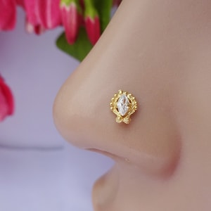 Puede incluir: Un pendiente de nariz de oro con una piedra preciosa transparente de corte marquesa en el centro, rodeada por un marco decorativo de cuentas. La pieza de joyería tiene un borde de metal texturizado y ornamentado que rodea la piedra.