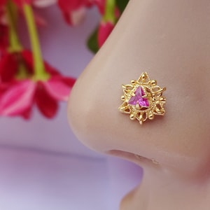 Puede incluir: Un pendiente de nariz de oro con un marco de metal intrincado de estilo filigrana y una piedra preciosa rosa de forma triangular en el centro. El trabajo decorativo del metal rodea la piedra en un patrón de estrella, creando un aspecto detallado.