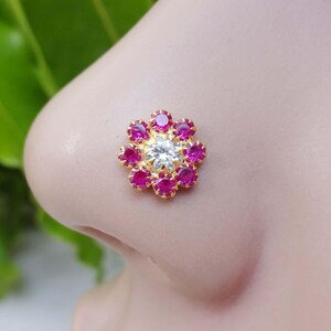 Könnte beinhalten: Ein goldener Nasenring mit einem rosafarbenen Blumendesign. Die Blume hat einen weißen Mittel-Stein und ist von kleineren rosafarbenen Steinen umgeben.